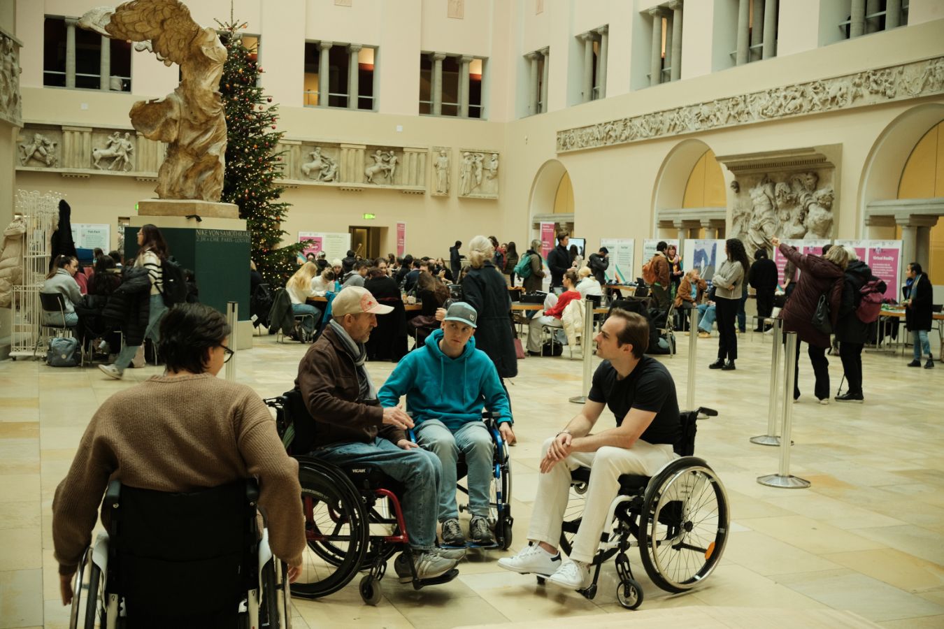 Rollstuhlsimulation: Mehrere Menschen, einige davon im Rollstuhl, sitzen und sprechen miteinander im Lichthof der UZH mit Informationsständen im Hintergrund.